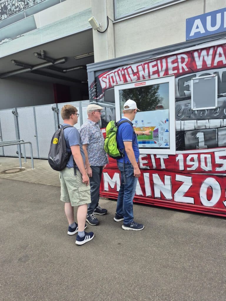 Drei Männer stehen vor einem Stadionplan, der an einer Wand mit Graffiti-artigen Motiven angebracht ist.
