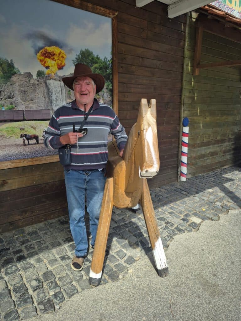 Ein Mann mit Cowboyhut posiert mit einem Holzpferd vor einem rustikalen Gebäude mit Steinpflaster.