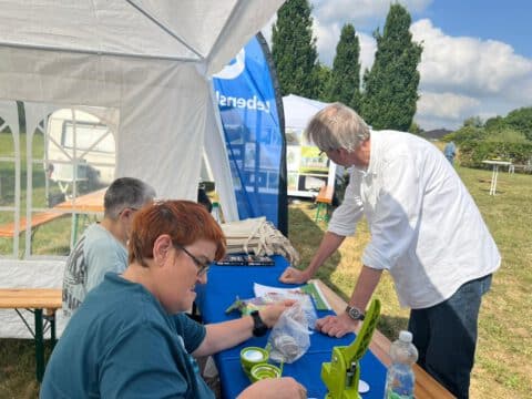 An einem sonnigen Tag arbeiten drei Personen an einem Tisch im Freien unter einem Vordach.
