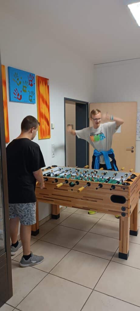 Zwei Personen spielen Tischfußball in einem hell erleuchteten Raum mit bunten Kunstwerken an der Wand.
