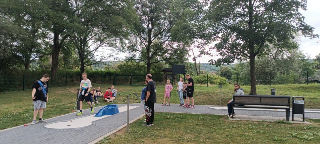 Eine Gruppe von Menschen spielt im Freien Minigolf, während andere zusehen oder auf einer Bank in der Nähe sitzen.