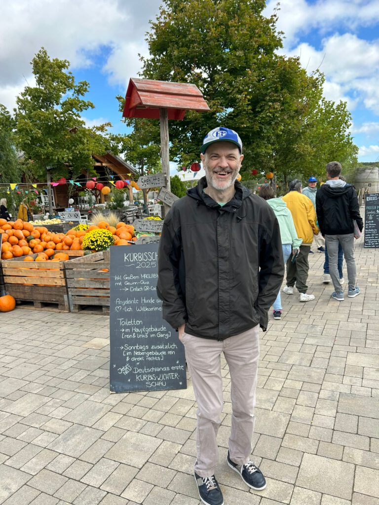 Ein Mann in einer schwarzen Jacke steht lächelnd auf einem Markt im Freien, auf dem Kürbisse und Schilder aus Kreidetafeln zu sehen sind.
