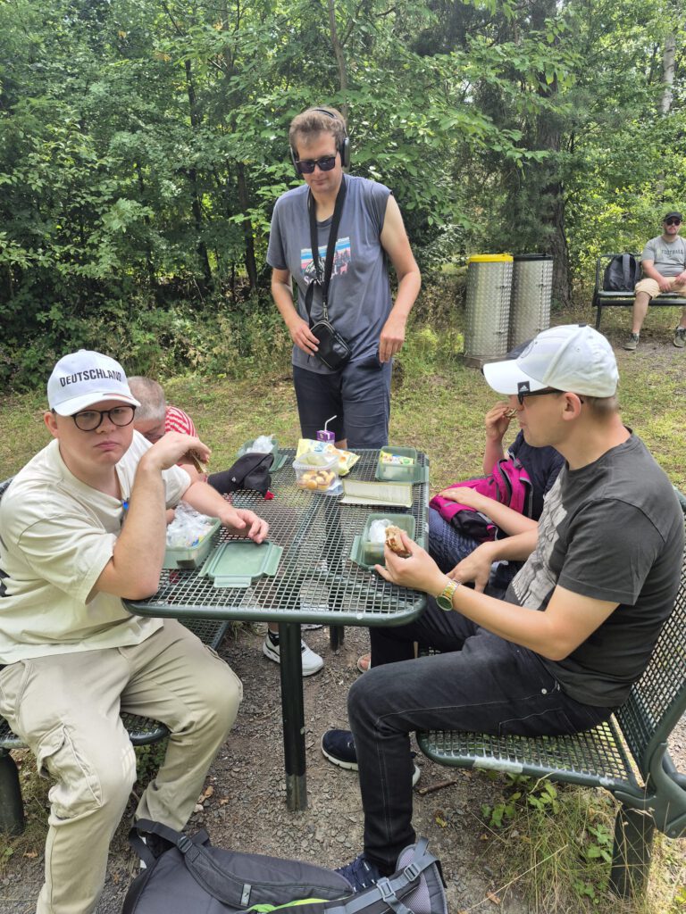 Vier Personen sitzen an einem Picknicktisch im Freien und essen zu Mittag; eine Person steht hinter dem Tisch.