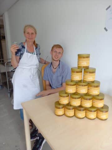 Zwei Personen mit einer Pyramide aus Honiggläsern auf einem Tisch in einem schlichten Innenraum.