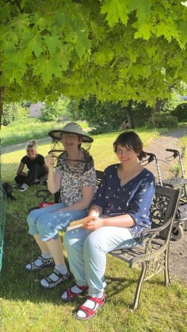 Zwei Frauen sitzen auf einer Bank unter einem Baum, eine weitere Person sitzt in der Nähe im Gras.