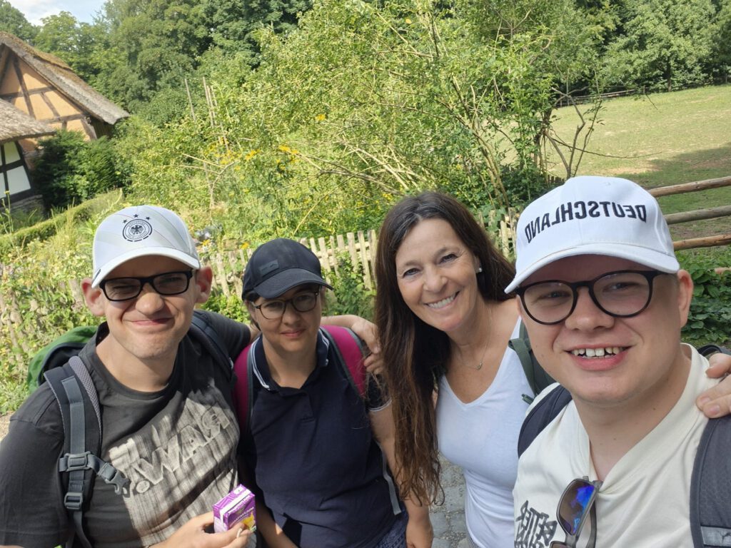 Vier Menschen stehen lächelnd im Freien, im Hintergrund sind Bäume und ein Holzzaun zu sehen.