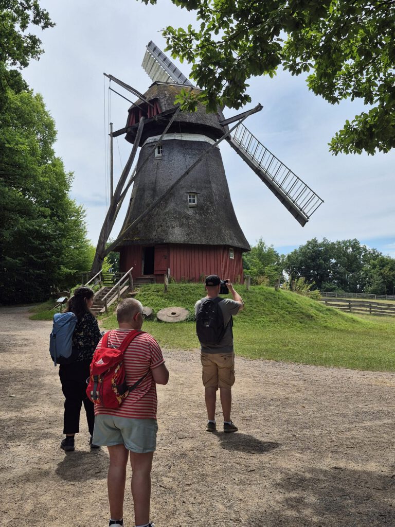 An einem sonnigen Tag stehen drei Menschen in der Nähe einer alten hölzernen Windmühle, die von Bäumen umgeben ist.