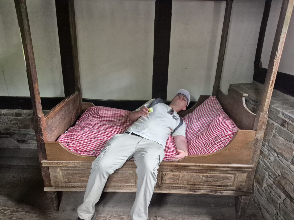 Eine Person liegt auf einem kleinen Holzbett mit rot karierter Bettwäsche und hält einen Gegenstand in der Hand.