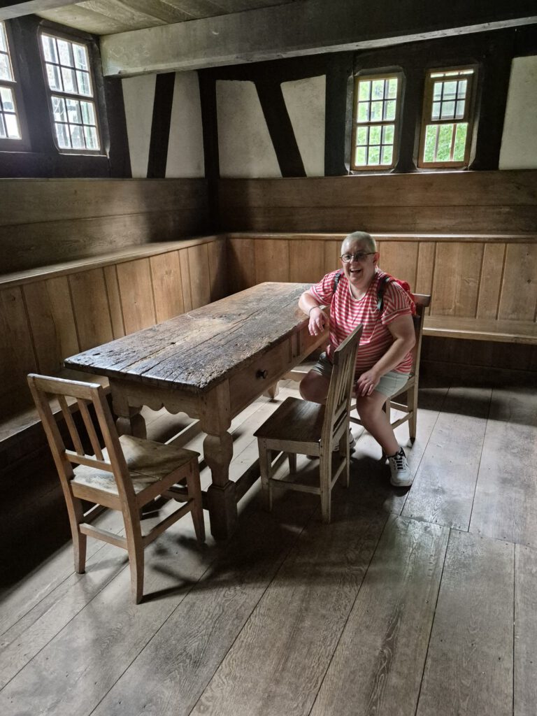Eine Person in einem gestreiften Hemd sitzt an einem rustikalen Holztisch in einem historischen Raum mit Fachwerkdecken.