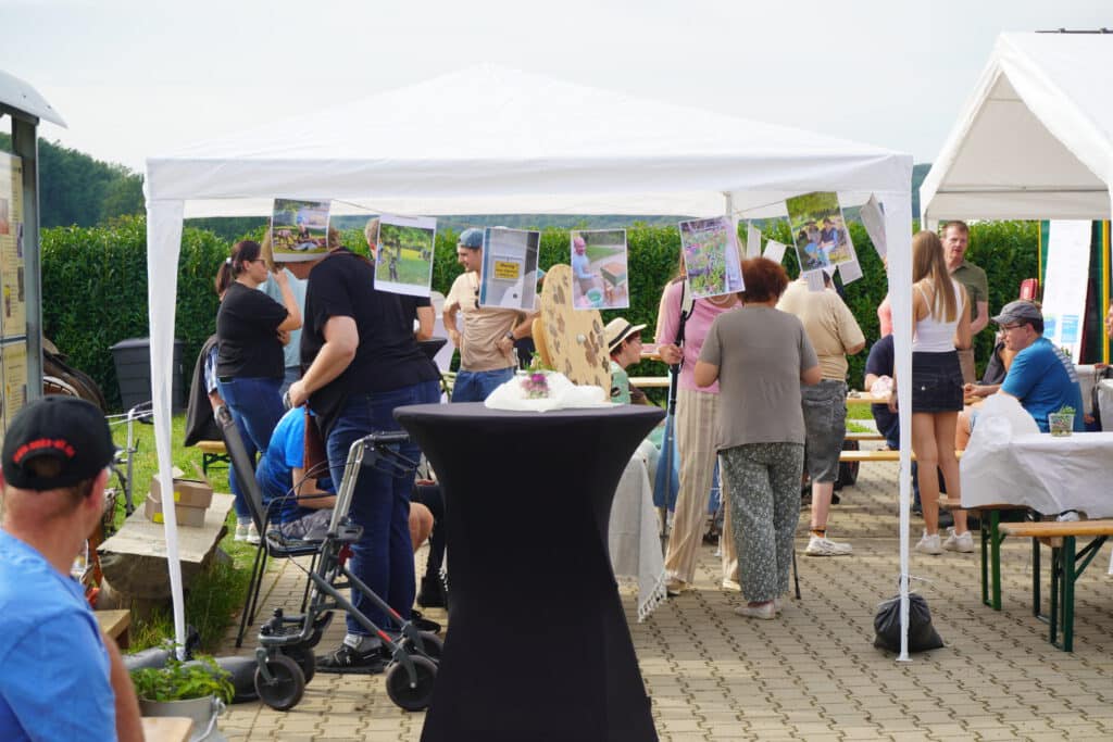 Unter einem weißen Zelt, an dem Plakate angebracht sind, versammeln sich Menschen, um an einer Veranstaltung im Freien teilzunehmen.