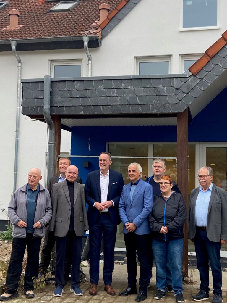 Sieben Männer und eine Frau stehen vor einem Gebäude und posieren für ein Gruppenfoto.