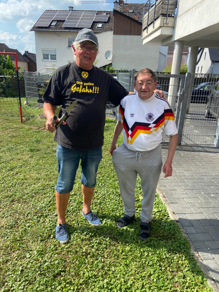 Zwei Männer stehen draußen im Gras, einer hält eine Zange, beide lächeln in die Kamera.