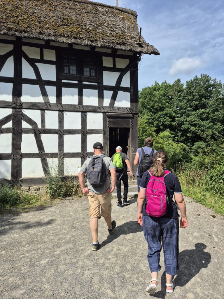 Vier Personen mit Rucksäcken gehen an einem sonnigen Tag auf ein Fachwerkhaus zu.