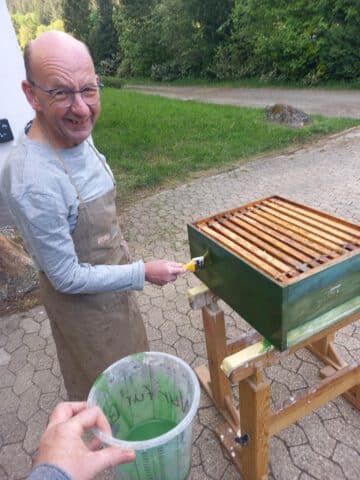 Ein Mann malt einen hölzernen Bienenkasten im Freien grün an, er hält einen Pinsel und trägt eine Schürze.