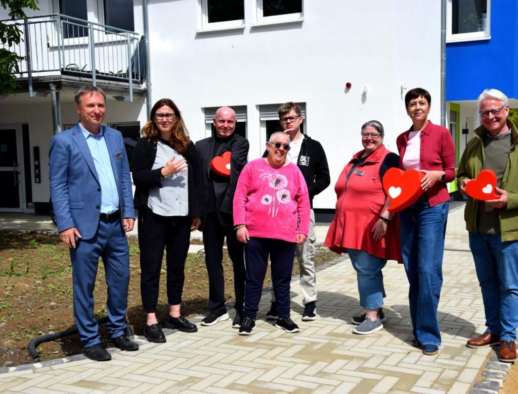 Acht Personen stehen vor einem Gebäude, einige halten rote, herzförmige Gegenstände in der Hand und lächeln in die Kamera.
