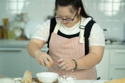 Eine Person in einer Schürze schlägt ein Ei in eine Schüssel, während sie in der Küche Essen zubereitet.