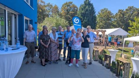 Eine Gruppe älterer Menschen posiert gemeinsam im Freien in der Nähe eines blauen Gebäudes während einer Veranstaltung bei sonnigem Wetter.