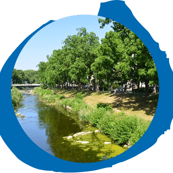 Ein Fluss, gesäumt von grünen Bäumen und Gras, fließt unter einem klaren blauen Himmel. In der Ferne ist eine weiße Brücke zu sehen. Das Bild wird von einem blauen kreisförmigen Grafikrahmen eingerahmt.
