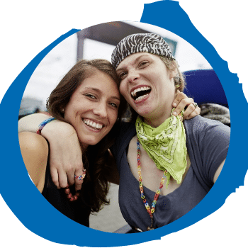 Zwei Menschen lächeln und posieren eng beieinander für ein Selfie im Freien, einer mit gemustertem Stirnband und grünem Bandana, der andere mit langen braunen Haaren, beide wirken glücklich und fröhlich.