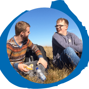 Zwei junge Männer sitzen unter einem klaren blauen Himmel auf trockenem Gras, lächeln und unterhalten sich. Einer hält ein Sandwich und eine Wasserflasche in der Hand, der andere trägt eine Brille und einen Kapuzenpullover. Beide wirken entspannt und glücklich im Freien.