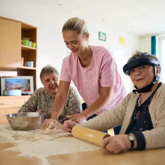 Eine Pflegekraft hilft zwei älteren Frauen beim Teigmachen am Küchentisch. Eine Frau rollt den Teig mit einer Nadel aus, die andere lächelt, und Mehl ist auf dem Tisch verteilt. Eine Rührschüssel und Küchenschränke sind zu sehen.